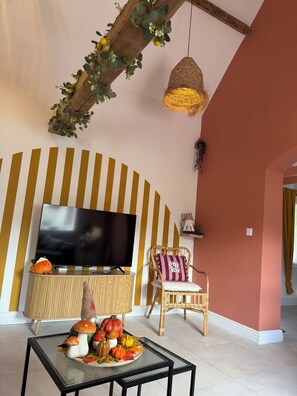 A living room with a television, a round table with decorative items, a wicker chair, and a striped wall. - Maison la Rosie - Air conditioning on the patio and in the garage (Blendecques)