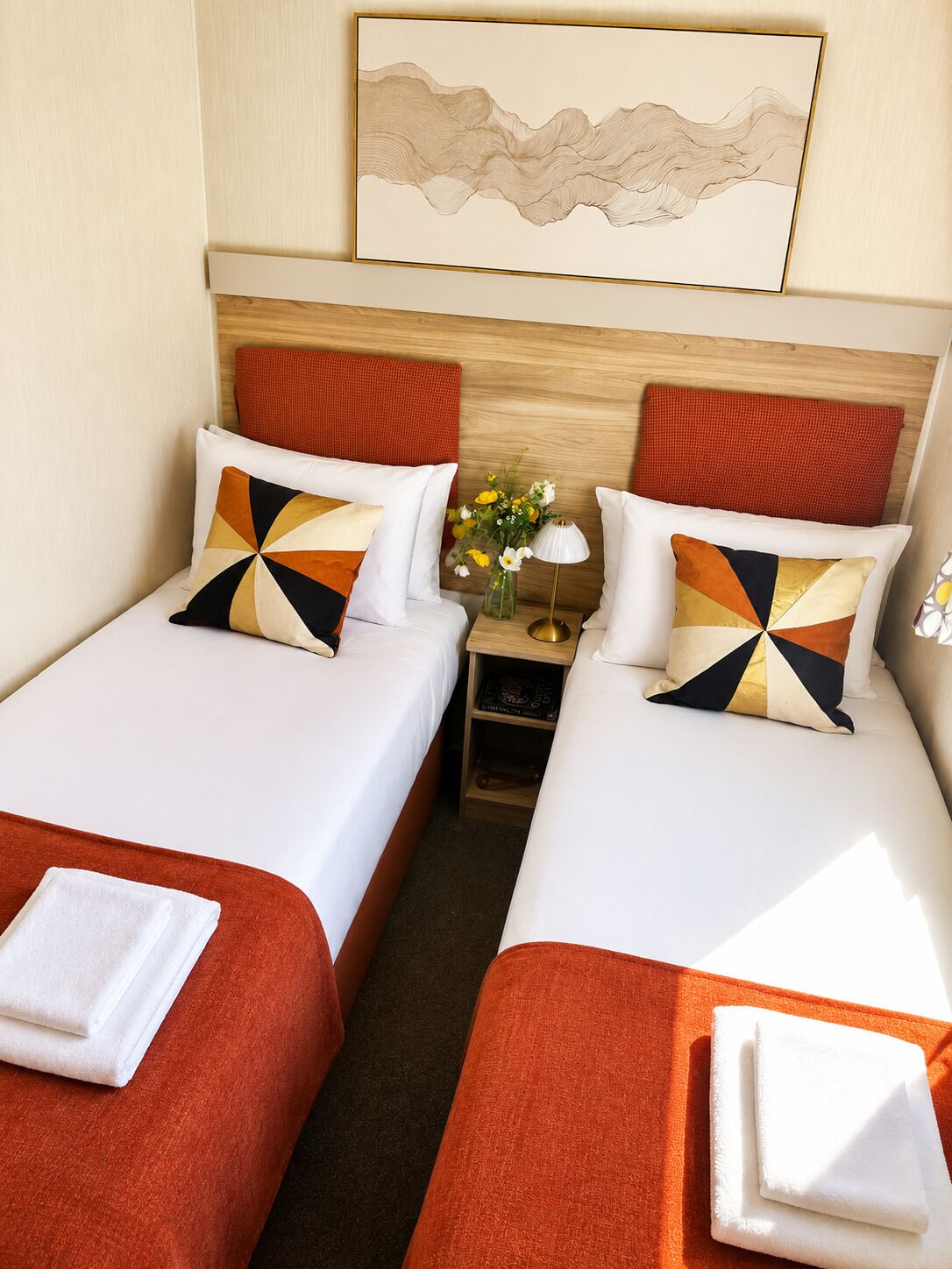 A hotel room with two beds, each with a red headboard, white bedding, and a geometric patterned pillow. There is a nightstand with a lamp and a vase of flowers between the beds.