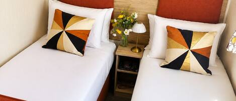 A hotel room with two beds, each with a red headboard, white bedding, and a geometric patterned pillow. There is a nightstand with a lamp and a vase of flowers between the beds.