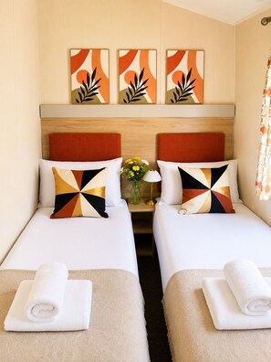 Ein Hotelzimmer mit zwei Betten, jeweils mit einem Kopfkissen und einem zusammengerollten Handtuch. Darüber hängen drei Bilder an der Wand.