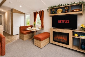 A cozy living area with a fireplace, a television displaying 'NETFLIX,' and a dining table set for two. - Anna by the Sea 3-Bed Sea Views & Blue Flag Beach (Devon)
