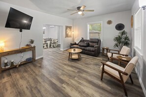 Ein Wohnzimmer mit Flachbildfernseher, Deckenventilator, Essbereich mit Tisch und Stühlen sowie ein großes Fenster mit Jalousien.