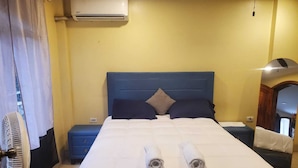 Une chambre d’hôtel comprenant un lit, deux tables de chevet, un appareil de climatisation, un ventilateur et une porte dotée d’un miroir.