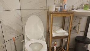 Une salle de bain comprenant des toilettes, une étagère en bois avec une serviette, une poubelle et un lavabo avec un distributeur de savon.