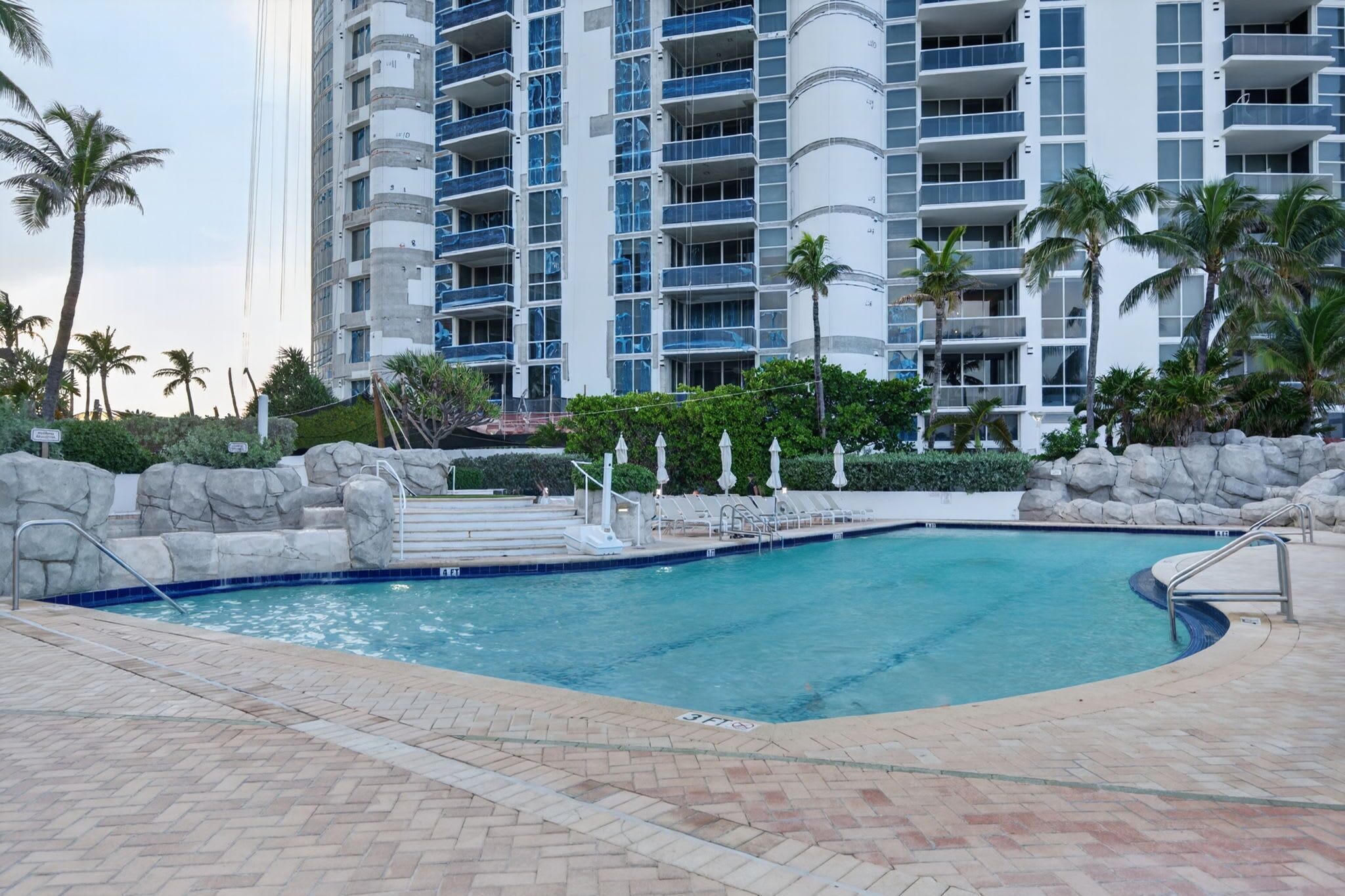 A swimming pool with a stone wall and palm trees, surrounded by a multi-story building with balconies.
