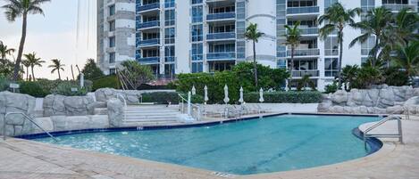 A swimming pool with a stone wall and palm trees, surrounded by a multi-story building with balconies.