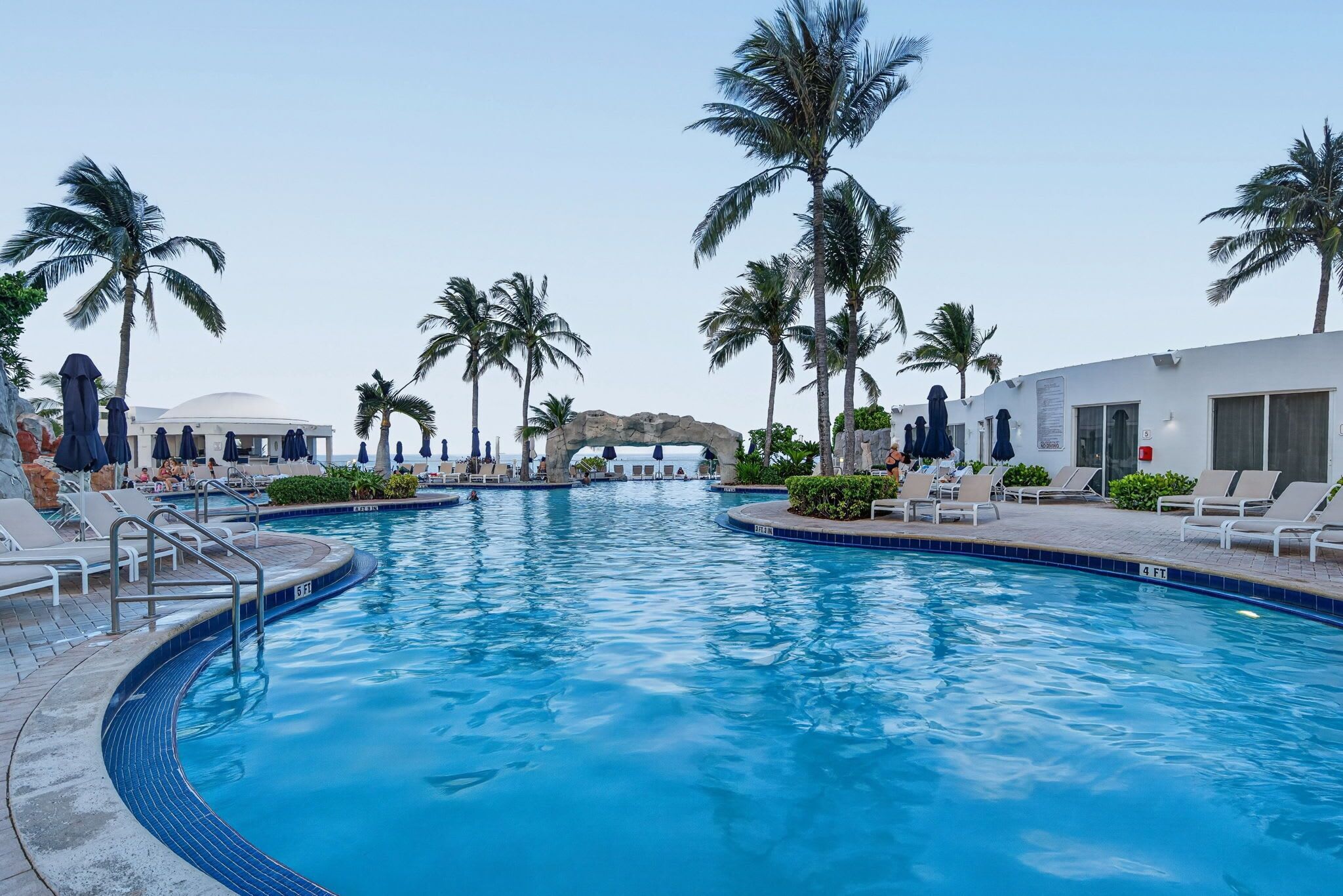A swimming pool with lounge chairs and umbrellas, surrounded by palm trees and a white building.