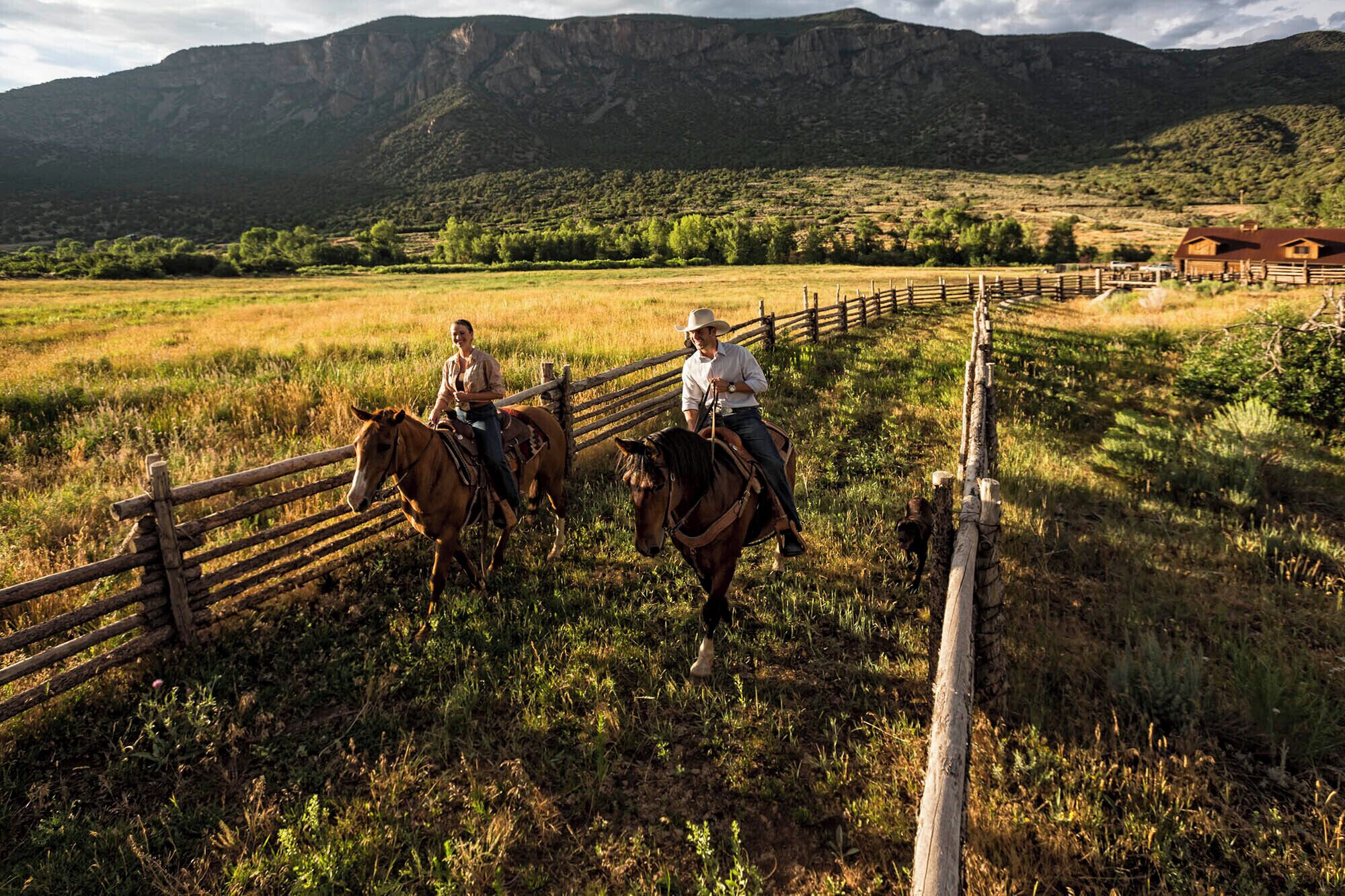 Two individuals riding horses along a wooden fence in a grassy field with mountains in the background.