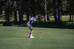 Eine Person spielt Golf auf einem gepflegten Rasen, im Hintergrund Bäume.