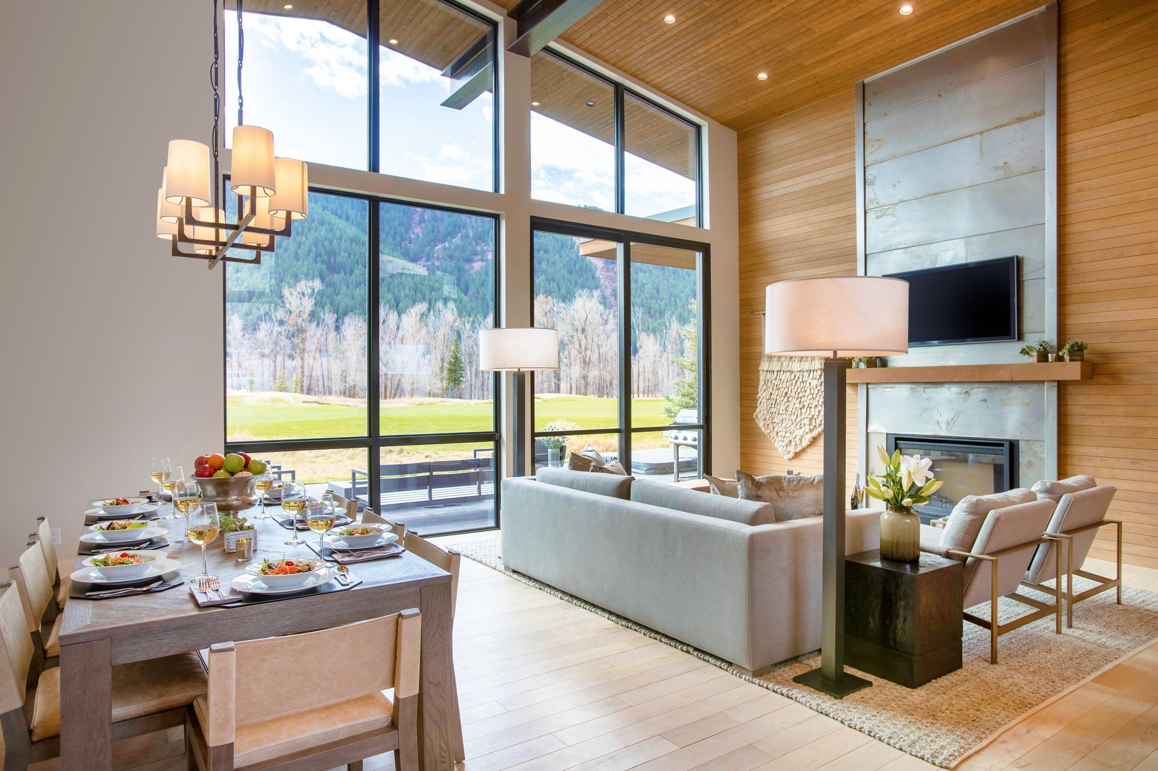 A modern dining area with a large table set for a meal, a fireplace, and a sofa in a room with large windows overlooking a forested landscape.