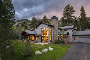 A modern two-story house with large windows and a stone chimney, surrounded by a well-maintained lawn and trees. - Luxury Private Mountain Home w/ Hot Tub (Jackson Hole)