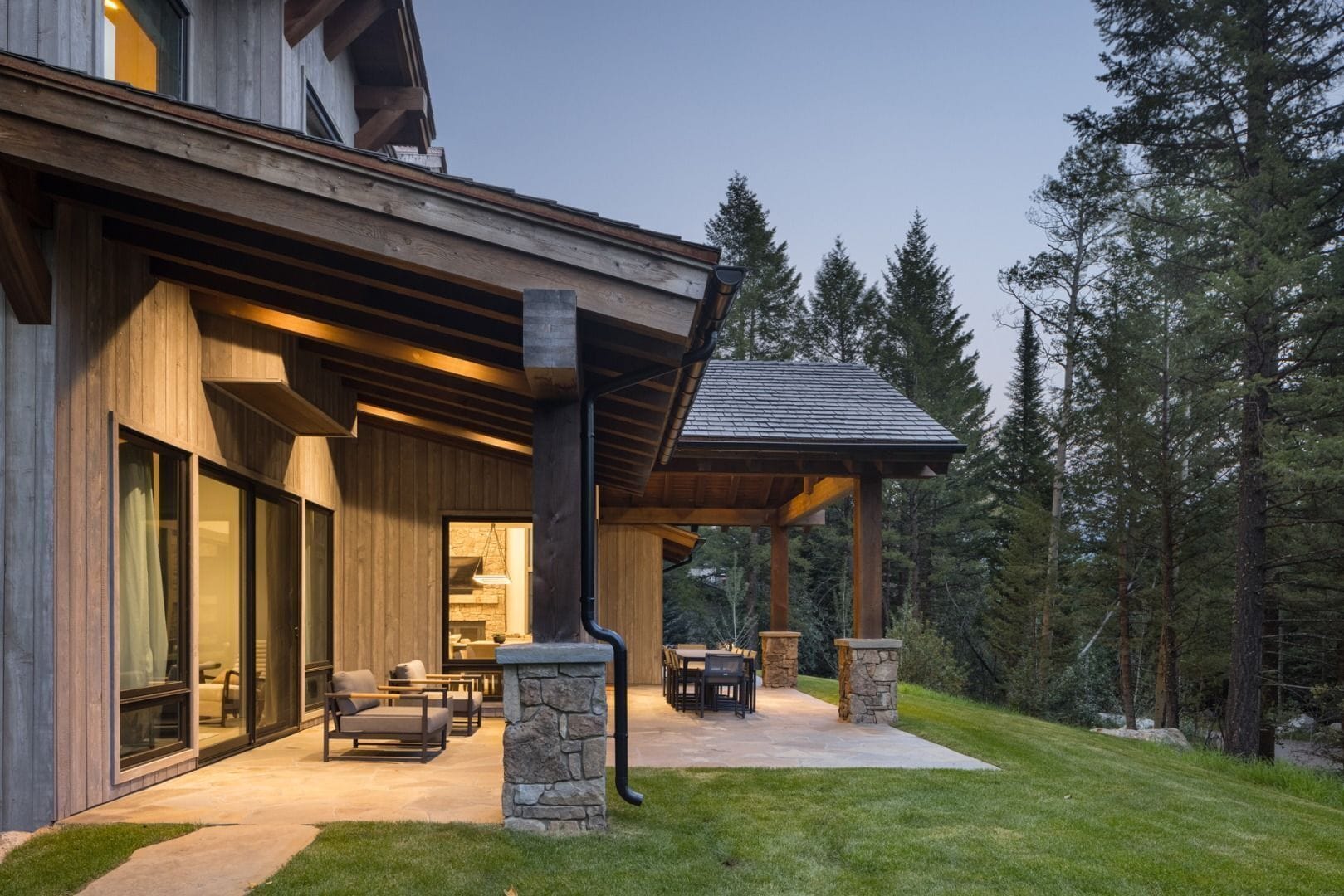 Ein modernes Haus mit einer überdachten Terrasse, Steinpfeilern und Blick auf den umliegenden Wald.