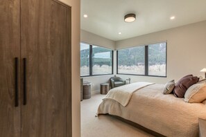 Ein Schlafzimmer mit Bett, Stuhl, Schreibtisch und Blick auf einen Wald durch große Fenster.