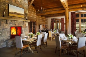 A dining area with wooden tables and chairs, a stone fireplace with a fire, and a large window with curtains. - Premier 3BR Fairway Cabin w/ Golf Views + Fire Pit (Jackson)