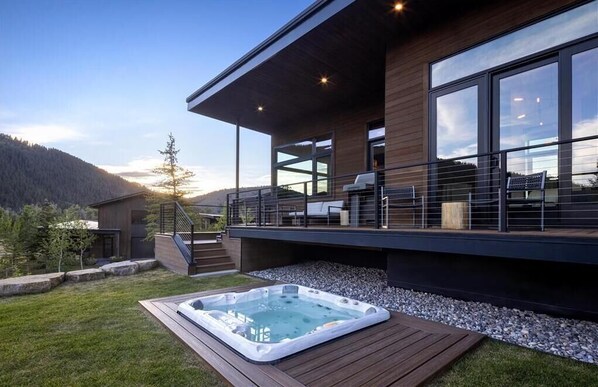 Cabin exterior with a deck and hot tub set against a densely forested hillside