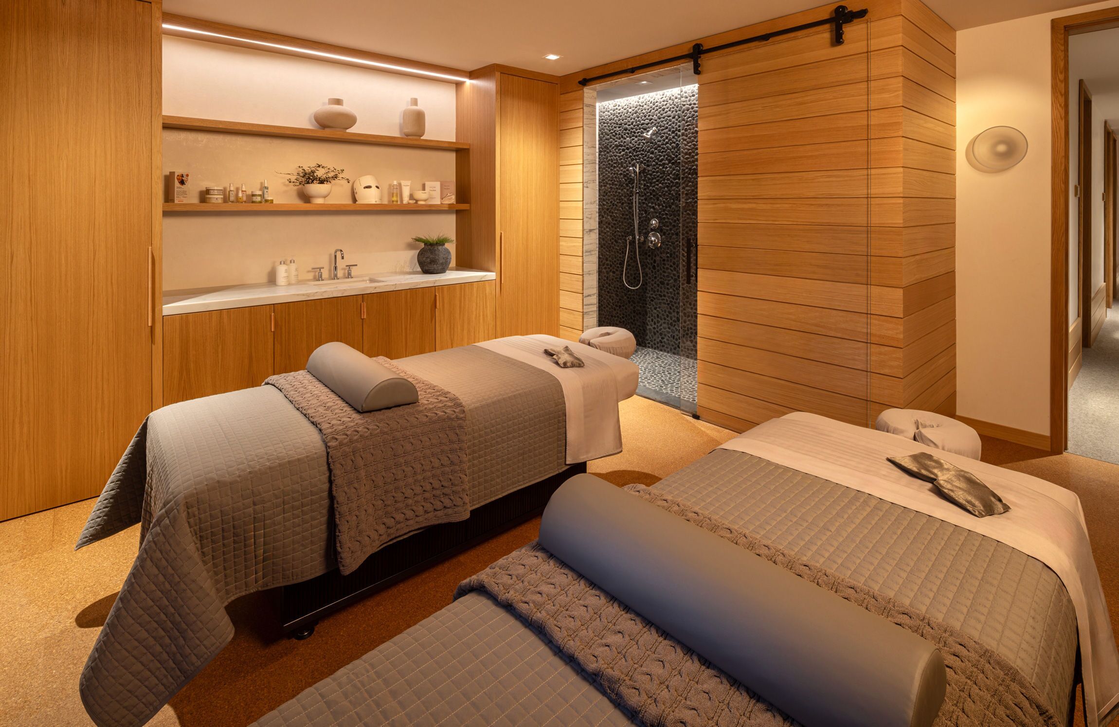 A spa room with two massage tables, a wooden wall, a shelf with toiletries, and a shower area.