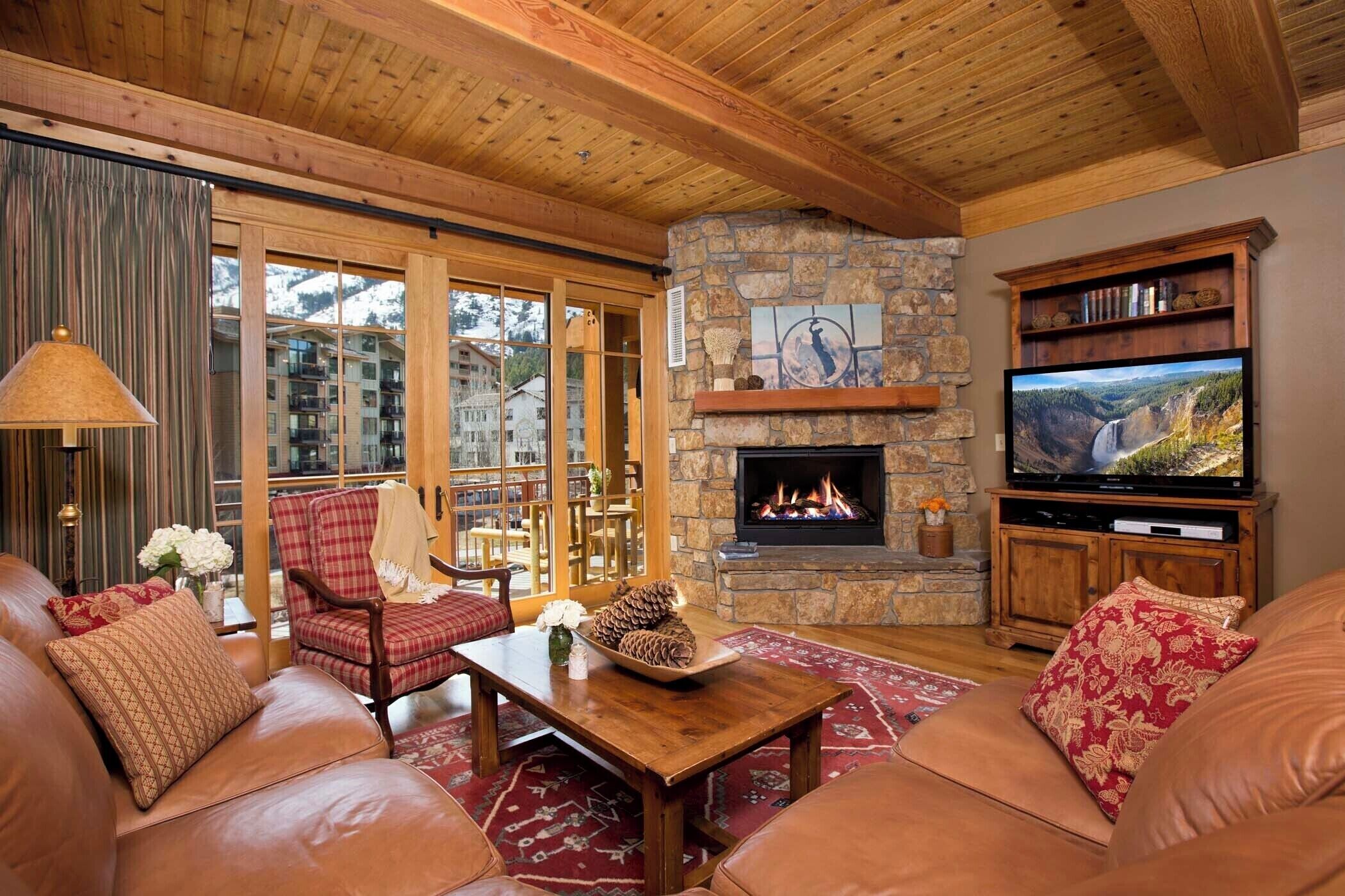 Ein gemütliches Wohnzimmer mit Ledersofas, einem Steinkamin, einem Fernseher und Blick auf eine gebirgige Landschaft durch große Fenster.