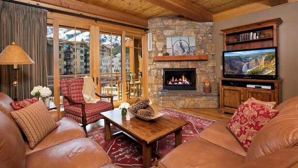 A cozy living room with leather sofas, a stone fireplace, a television, and a view of a mountainous landscape through large windows. - Ski-In Condo | Tram-Adjacent | Mountain Views (Jackson Hole)