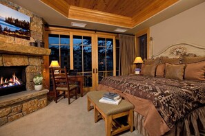A cozy bedroom with a stone fireplace, a bed with patterned bedding, and a view of the outdoors through large windows. - Ski-In Condo | Tram-Adjacent | Mountain Views (Jackson Hole)