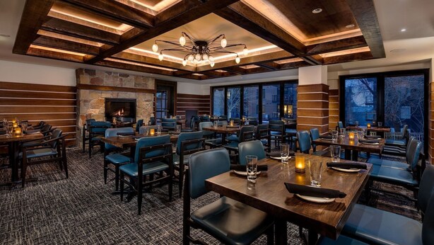 A dining area with multiple tables and chairs, a stone fireplace, and a view through large windows. - Slopeside Condo w/ Mountain Views | Ski-In Access (Jackson Hole)