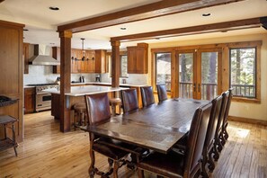 A spacious kitchen with wooden cabinets and a large island, a dining area with a wooden table and chairs, and large windows with a view of trees. - Grand Alpine Lodge w/ Sauna, Hot Tub & Game Room (Jackson Hole)