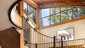 A wooden staircase with a curved design leading to a second-floor area with large windows and a bear painting. - Grand Alpine Lodge w/ Sauna, Hot Tub & Game Room (Jackson Hole)