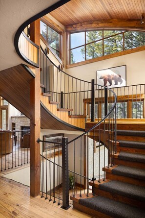 A wooden staircase with a curved design leading to a second-floor area with large windows and a bear painting. - Grand Alpine Lodge w/ Sauna, Hot Tub & Game Room (Jackson Hole)