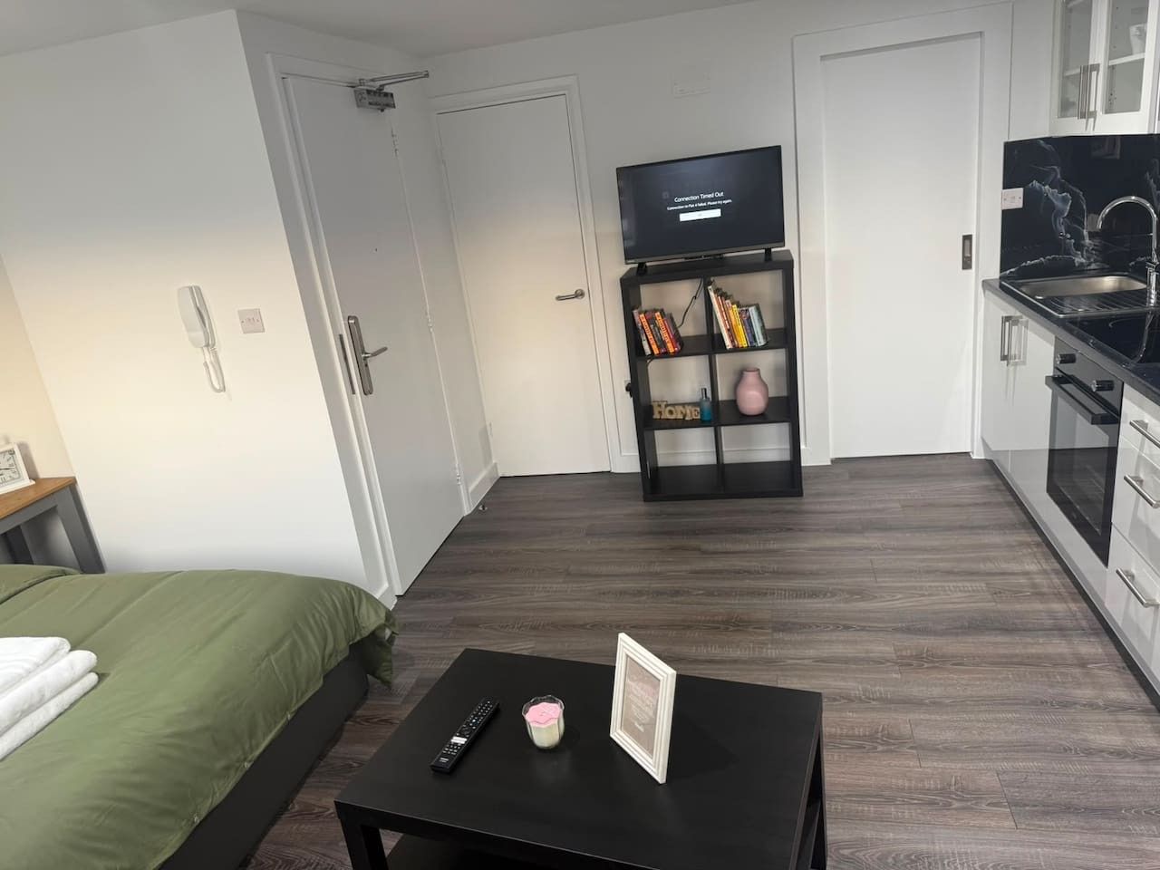 Ein Hotelzimmer mit einem Bett, einem kleinen Tisch mit Fernbedienung und Kerze, einem Fernseher auf einem Schrank und einer Küchenzeile mit Spüle und Herd.