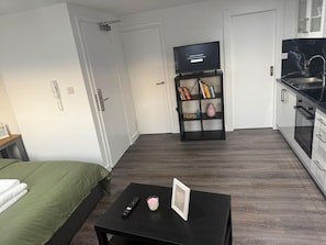 Ein Hotelzimmer mit einem Bett, einem kleinen Tisch mit Fernbedienung und Kerze, einem Fernseher auf einem Schrank und einer Küchenzeile mit Spüle und Herd.
