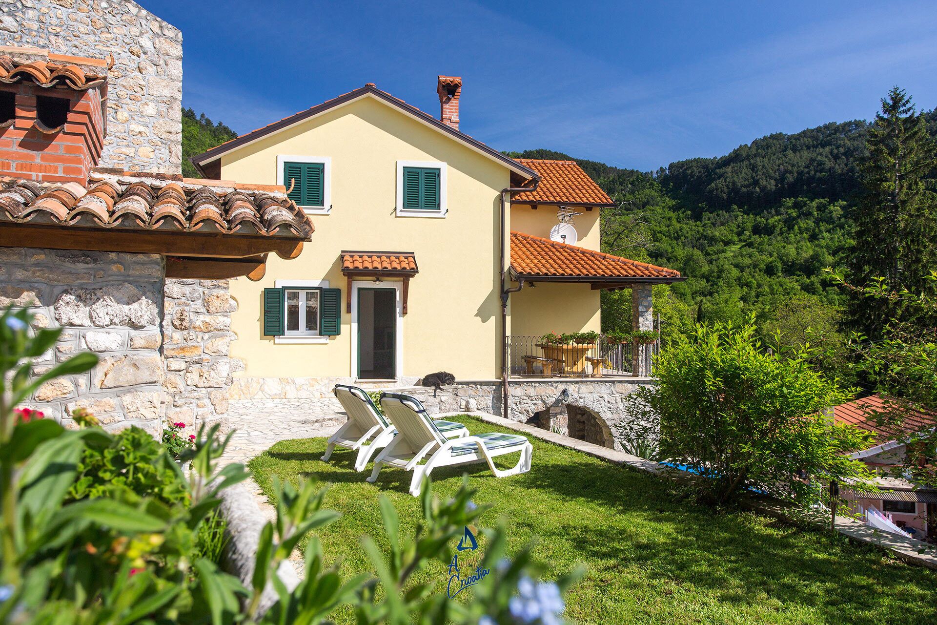 A two-story house with a stone facade, a tiled roof, and a small balcony with outdoor furniture.