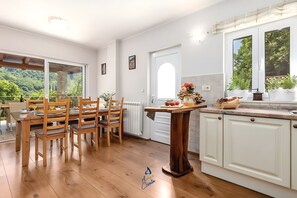 A kitchen with wooden cabinets, a dining table with chairs, a view of greenery outside, and a window with a view of trees. - Charming 5-BR Villa with Pool near Beaches (Lovran)