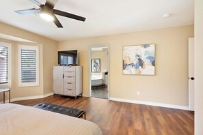 A bedroom with a bed, a dresser, a ceiling fan, a painting, and a bathroom visible through a door. - Cozy 3-bedroom home in beautiful Henderson with cool pool (Henderson)