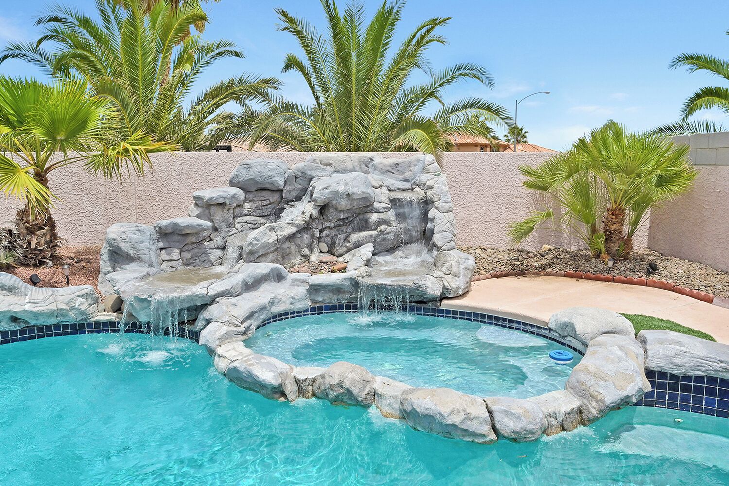 A swimming pool with a rock waterfall feature and palm trees in the background.