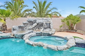 Piscina com cachoeira de pedra e palmeiras ao fundo.