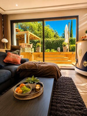 Ein modernes Wohnzimmer mit einer Couch, einem Couchtisch mit Obst und einer Kerze, sowie einer großen Schiebetür zu einem Außenbereich mit Holzterrasse und viel Grün.