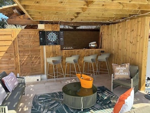 A wooden outdoor seating area with a dartboard, bar stools, and a table with a decorative bowl. - Villa with pool & pool house – AC – 6/8 guests (Roquebrune-sur-Argens)