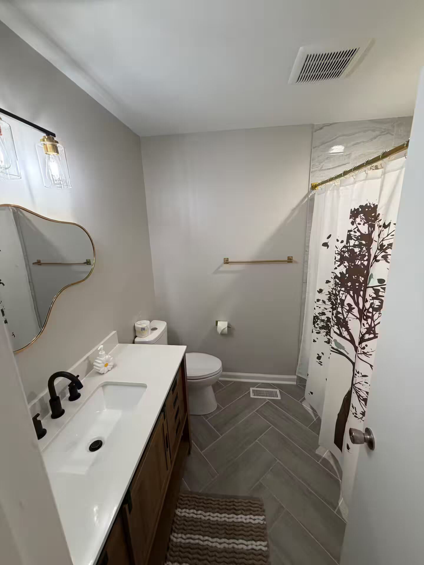 Une salle de bain avec un lavabo blanc, des toilettes, un rideau de douche orné d’un motif d’arbre et un miroir avec des luminaires.