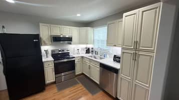 Cocina compacta con muebles blancos, un refrigerador negro, electrodomésticos de acero inoxidable y una ventana con persianas.