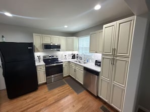 A compact kitchen with white cabinetry, a black refrigerator, stainless steel appliances, and a window with blinds.
