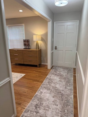 A hallway with a wooden floor, a gray rug, a wooden dresser, a lamp, and a white door. - Modern Comfort 3BR  Coliseum, Koury Convention, High Point Market (Greensboro)