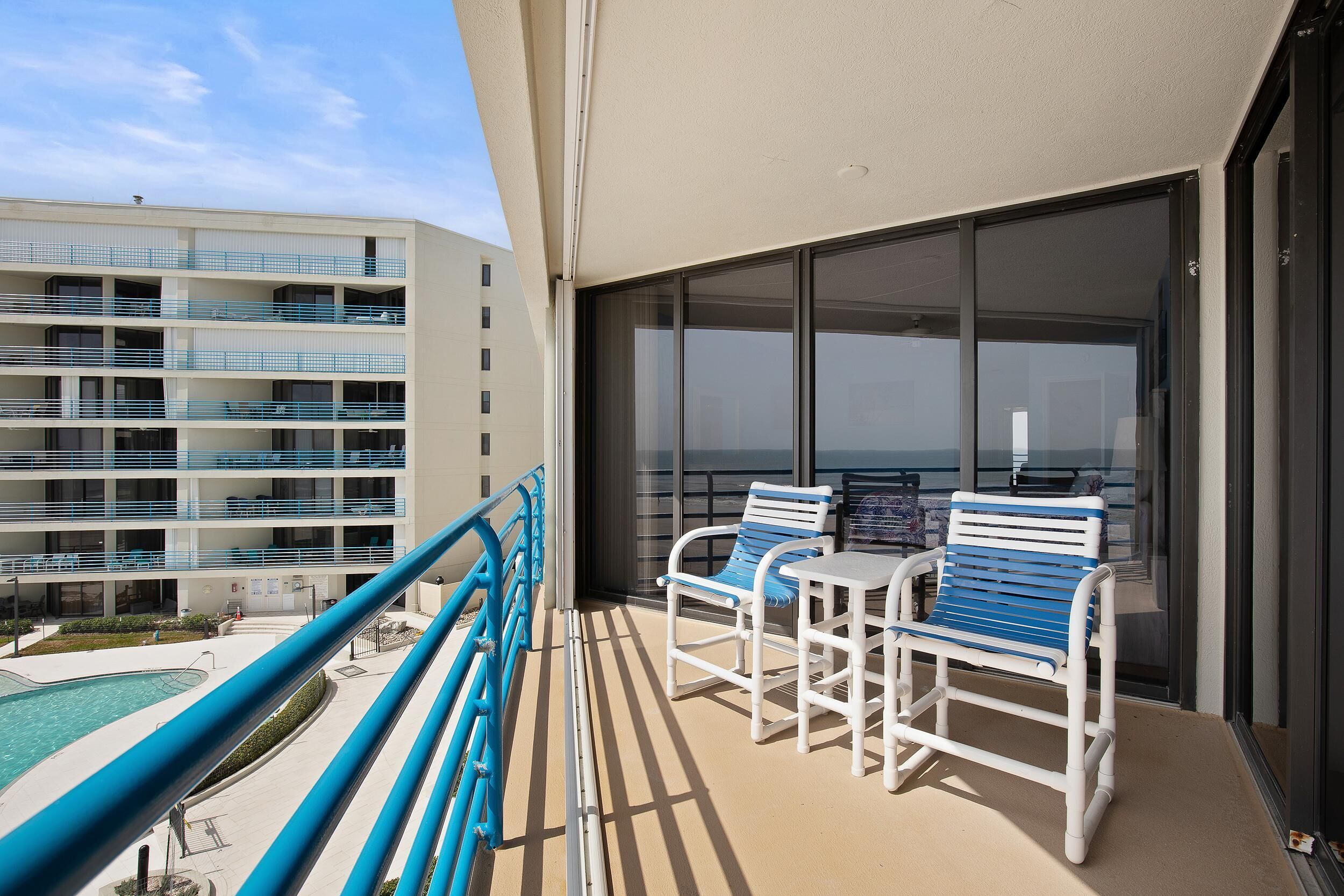 A balcony with two white chairs and a table, overlooking a pool and a building with multiple balconies.