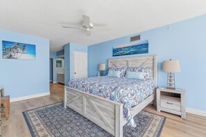A bedroom with a bed, two bedside tables, a ceiling fan, and artwork on the walls. - Ebb Tide 406 (New Smyrna Beach)