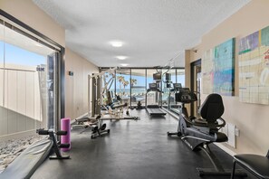 A gym with exercise equipment, a treadmill, and a large window overlooking a pool and palm trees. - Ebb Tide 406 (New Smyrna Beach)