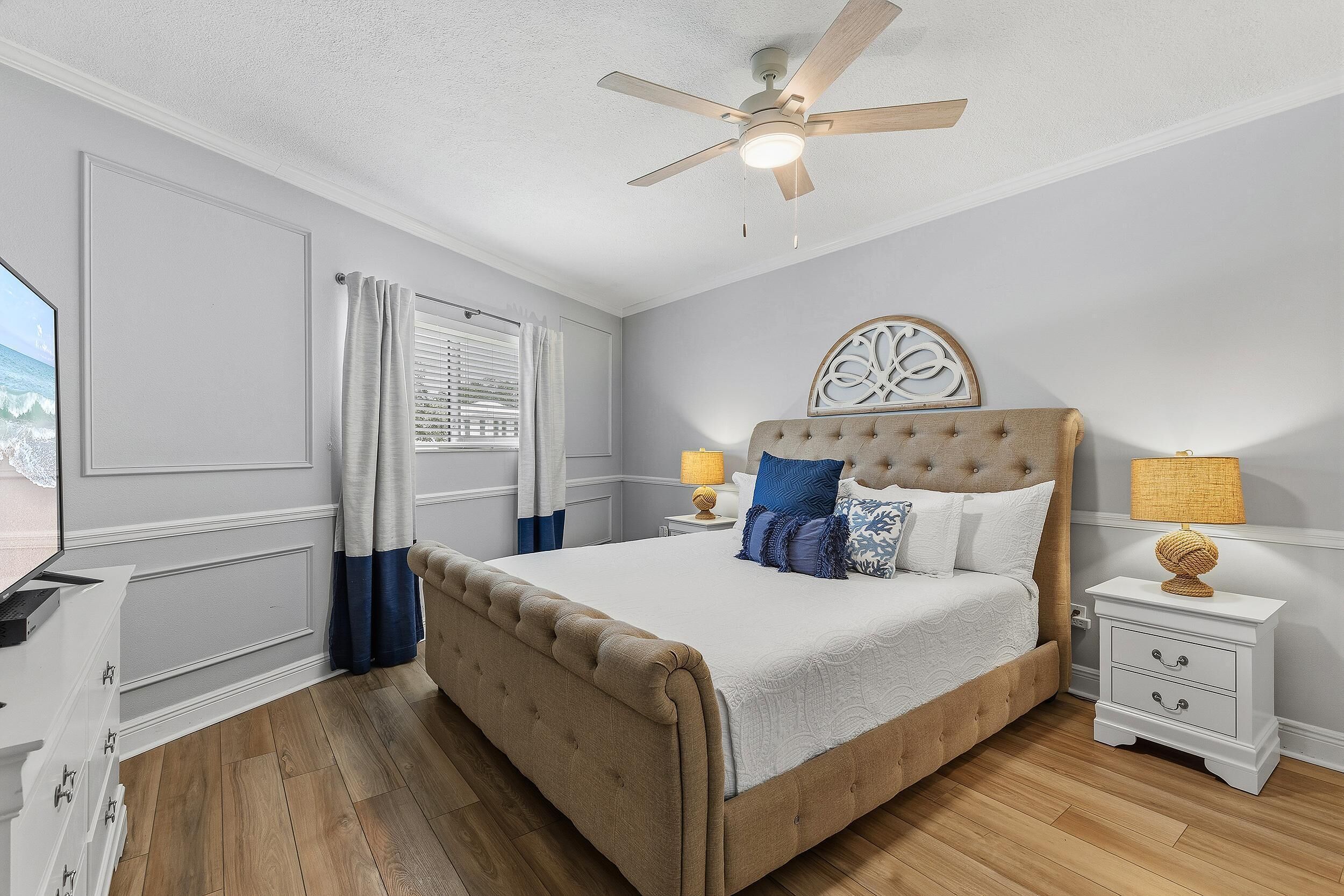 Une chambre à coucher avec un grand lit, un ventilateur de plafond, une table de chevet avec une lampe et une fenêtre avec des rideaux.