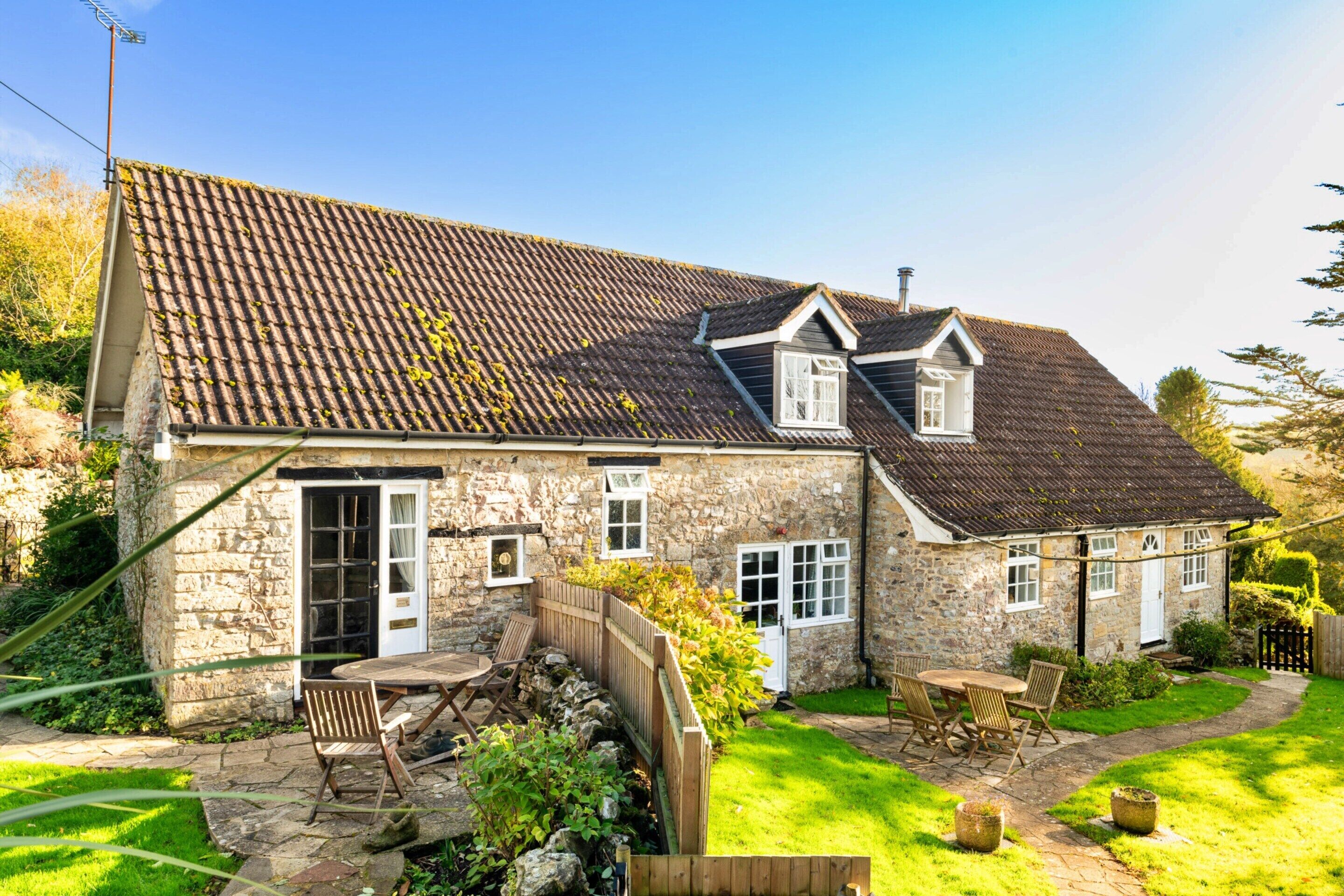 Ein Steinhhaus mit Ziegeldach, einem Holzzaun und einem Garten mit Terrassentisch und -stühlen.