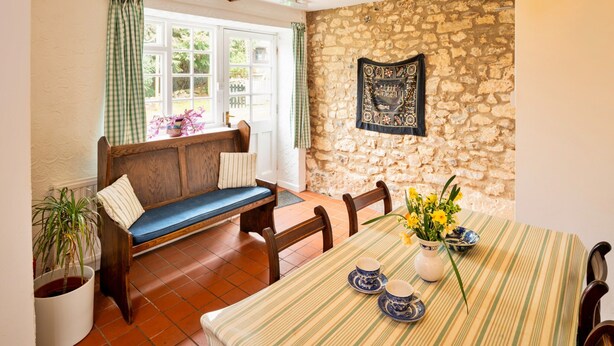 A cozy dining area with a stone wall, a bench, a table with a striped tablecloth, and a vase with flowers. - Coastal Family House for 8 in Cornwall (Axminster)