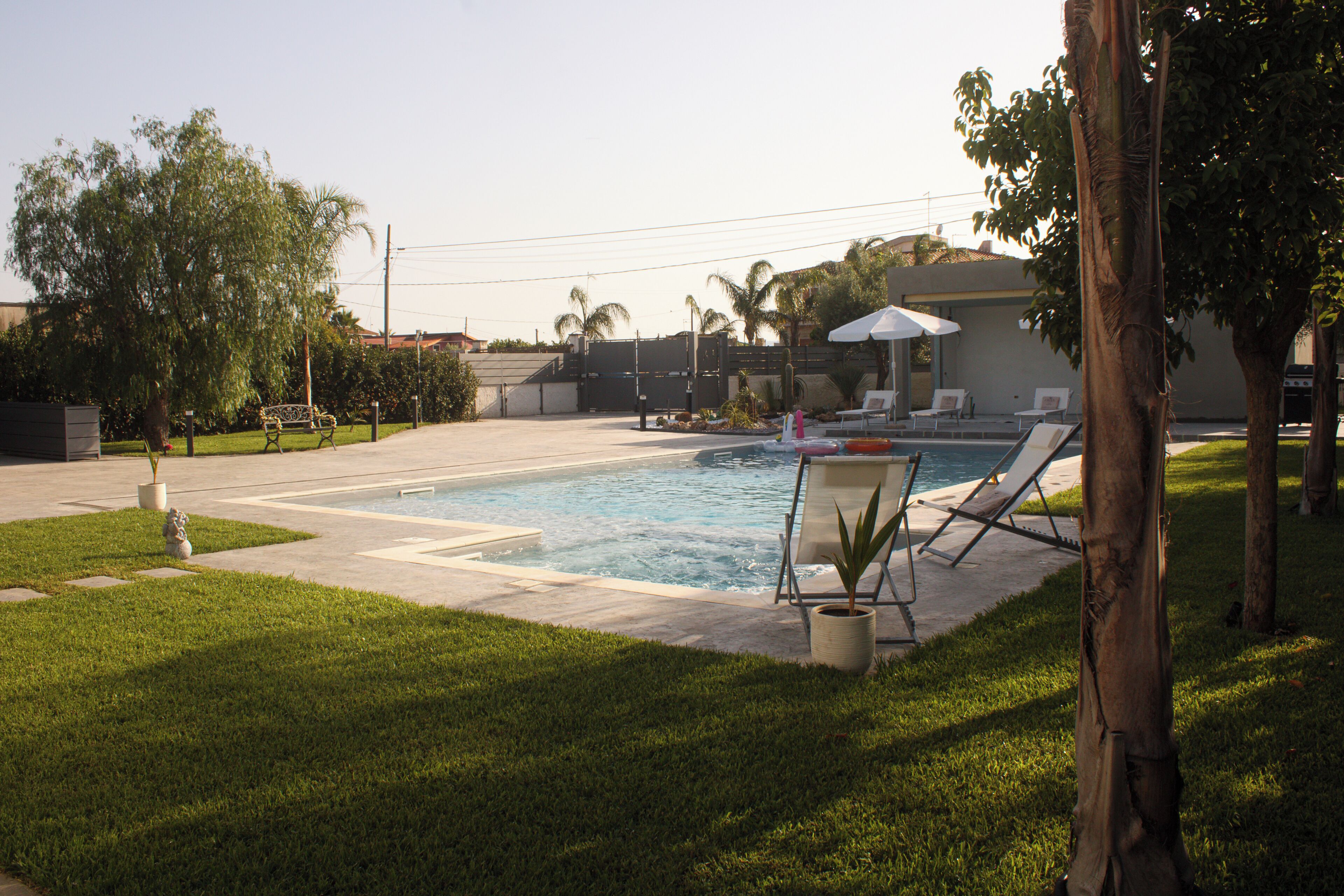 A swimming pool with lounge chairs and a covered area, surrounded by grass and trees.