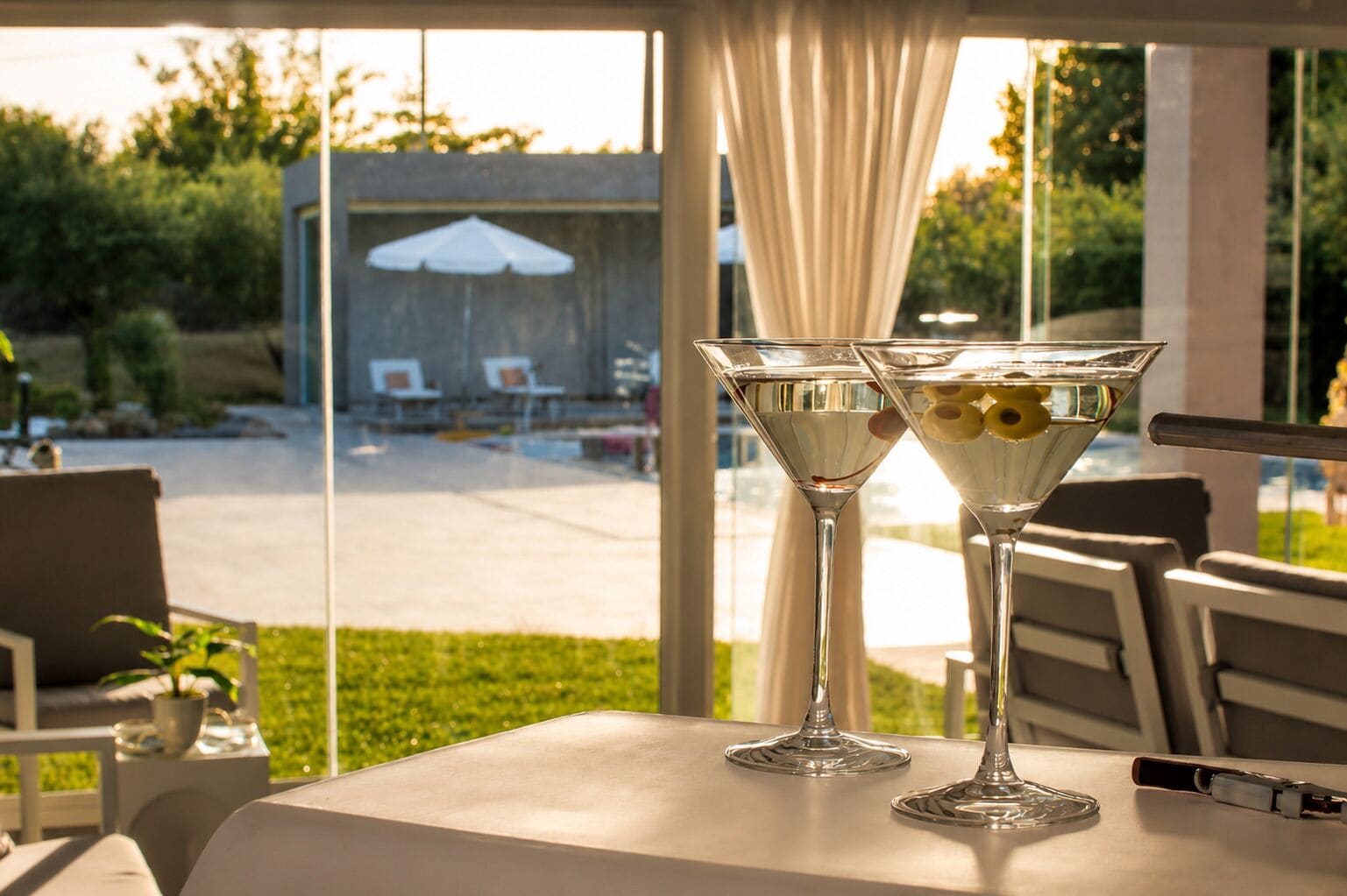 Une table avec deux verres à martini et une bouteille, placée devant une fenêtre donnant sur un patio aménagé avec du mobilier d’extérieur.