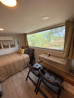 A compact room with a bed, a desk, and a chair. There is a window with a view of greenery outside.