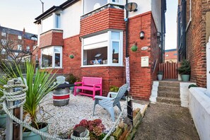 A residential property with a red brick exterior, a white window, and a small garden area with a pink bench and a white chair. - Welcoming Cottage Near Harbour (Whitby)
